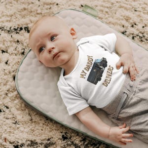 Peut inclure: Un body blanc pour bébé avec l'inscription "MY DADDY DELIVERS" en lettres dorées et un dessin d'un camion de livraison. Le bébé est allongé sur une surface rembourrée verte et grise.