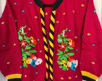 Può includere: Un cardigan di maglia rosa con un bordo intrecciato nero e giallo. Il cardigan presenta un motivo floreale con foglie verdi, fiori rossi, arancioni, gialli e blu.