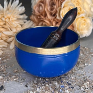 May include: A blue singing bowl with a gold rim and a dark wooden striker. The bowl is a vibrant blue colour, and the striker is dark brown. The bowl is sitting on a surface with small, shiny, reflective pieces.