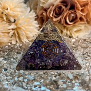 Könnte beinhalten: Eine orgon-Energiepyramide mit einer Basis aus lila Amethystkristallen und einer Kupferspirale. Die Pyramide hat goldfarbene Akzente und wird auf einer reflektierenden Oberfläche präsentiert. Verschwommener floraler Hintergrund.