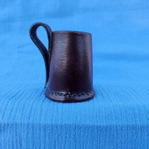 Peut inclure: Une tasse en cuir marron foncé avec une anse. La tasse est posée sur une surface en tissu bleu.