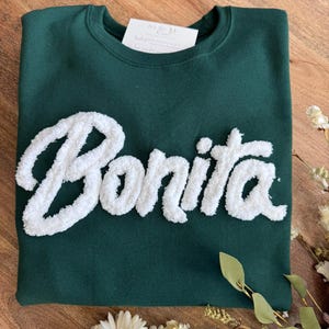 May include: A dark green crewneck sweatshirt featuring the word Bonita in large, white, textured, fuzzy chenille-style lettering across the chest. A small white rectangular tag is tucked inside the collar area of the garment.