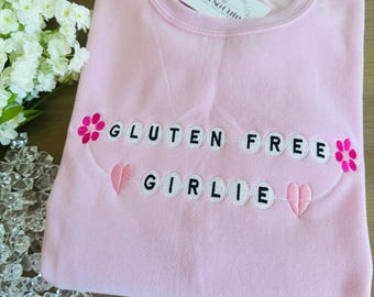 May include: A light pink crewneck sweatshirt featuring embroidered white circular patches with black lettering that reads GLUTEN FREE GIRLIE. The design includes two pink flowers and two pink hearts stitched onto the fabric of the garment.