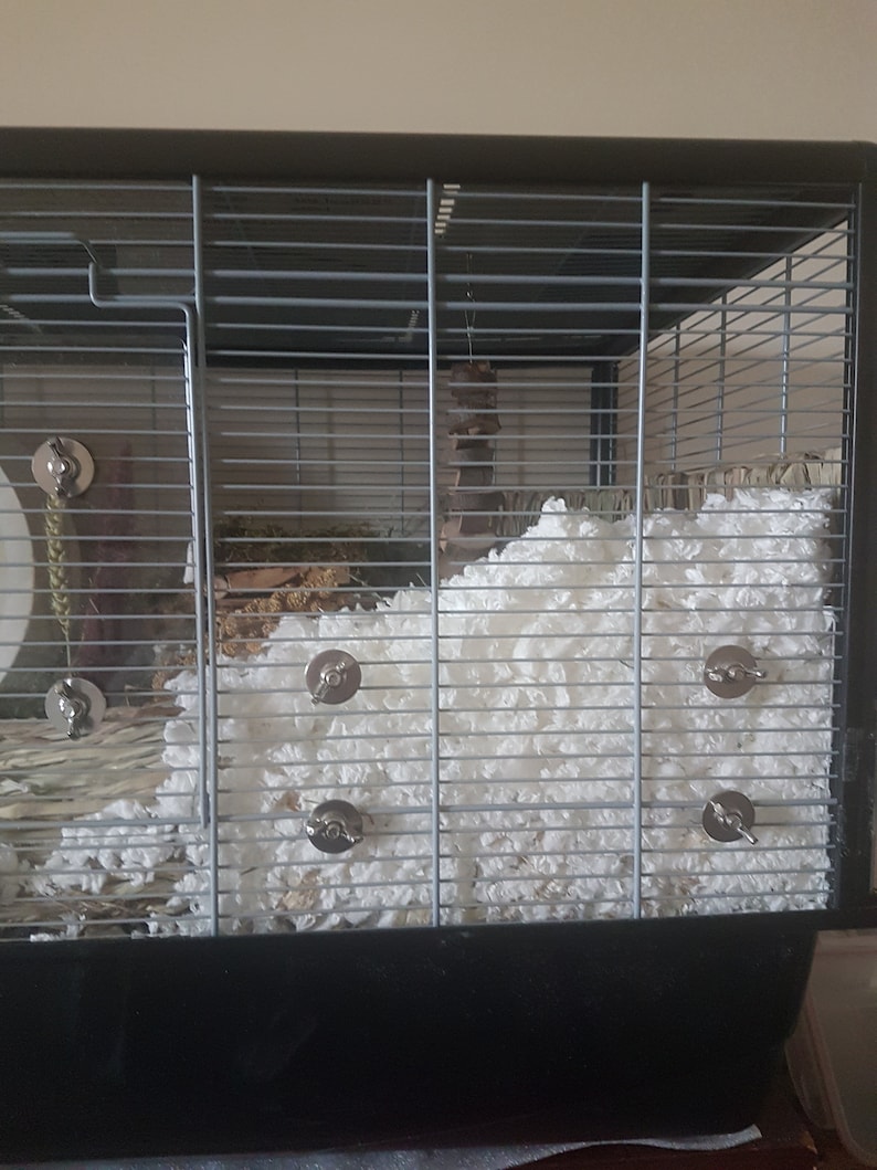 May include: A black metal cage with a white fluffy bedding material. The cage has a metal door and a metal latch. There are several metal clips on the side of the cage.