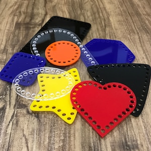 May include: Assortment of colourful plastic crochet stitch markers in various shapes, including a heart, star, circle, square, and rectangle. The markers are clear with a white outline and have holes around the edges.
