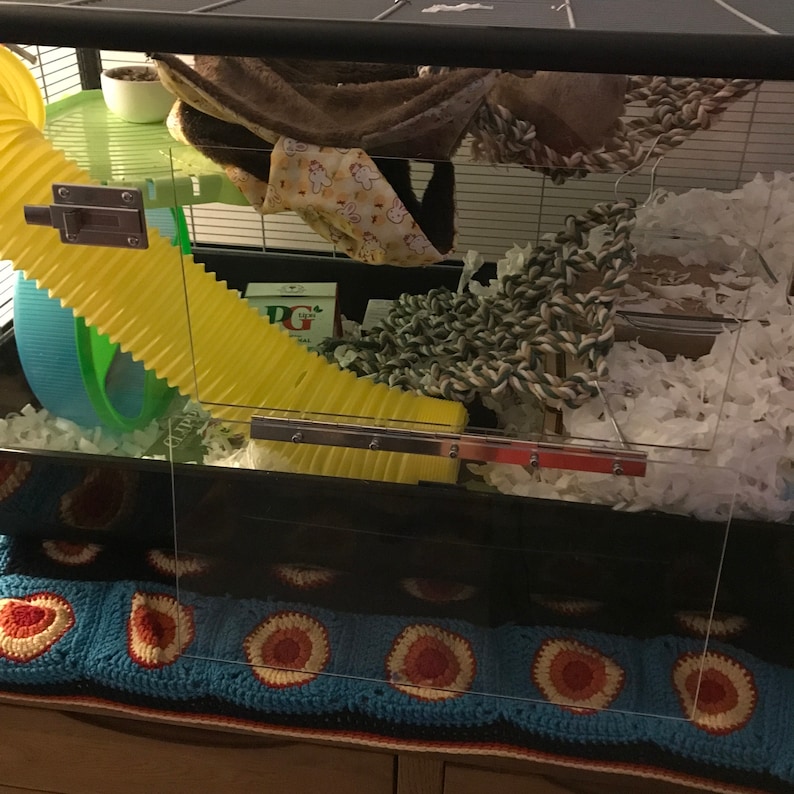 May include: A yellow plastic tube, a green and blue exercise wheel, and a brown and white fabric hammock are inside a clear plastic cage. The cage has a white and green rope toy, white shredded paper, and a small brown bowl. The cage is sitting on a blue and red crocheted blanket.