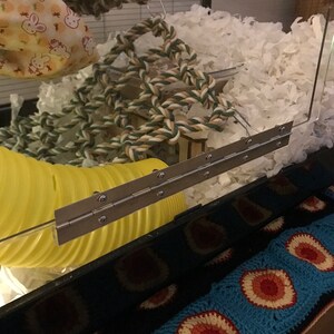 May include: A close-up of a silver metal hinge attached to a clear plastic cage. The hinge is attached to a yellow plastic tube and a wooden board. The cage is filled with white shredded paper.
