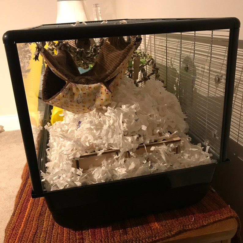 May include: A black wire cage with a brown and white hanging hammock, white shredded paper bedding, and two wooden platforms.