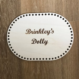 May include: A white oval wooden sign with black dots around the edge. The sign says "Brinkley's Dolly" in a brown cursive font.