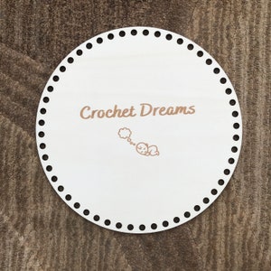 May include: White round crochet board with black dots around the edge. The board has the text "Crochet Dreams" written on it in brown lettering. A small cartoon of a sleeping baby with a thought bubble above its head is also on the board.
