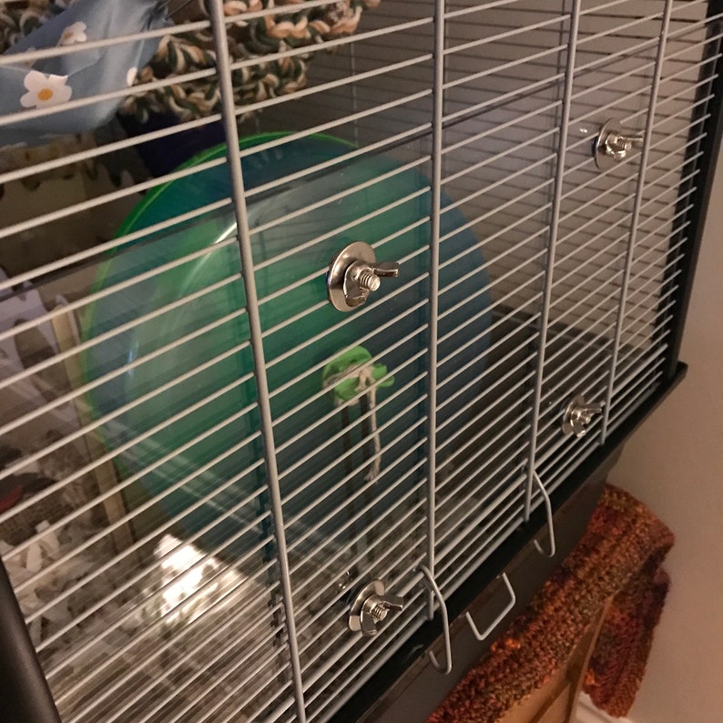 May include: A black metal hamster cage with a green plastic exercise wheel and multiple silver clips. The cage is sitting on a brown and orange patterned surface.