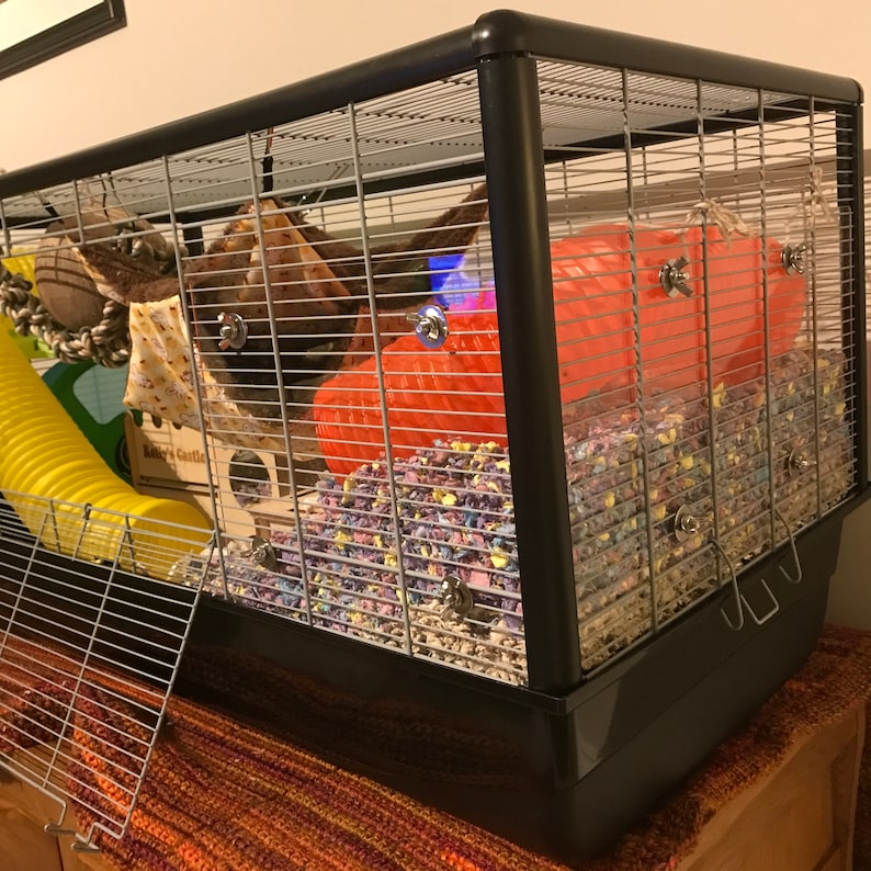 May include: A black wire cage with a red and white plastic tube, a yellow tube, and a wooden platform. The cage is filled with colorful bedding.