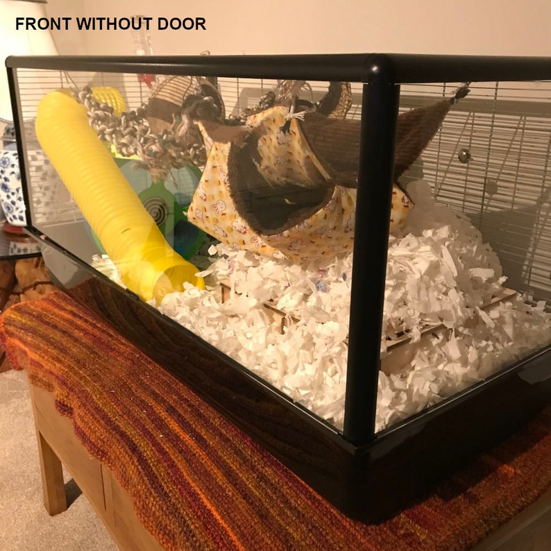 May include: A black hamster cage with a yellow tube, a brown and yellow hammock, and white bedding. The cage is labeled "FRONT WITHOUT DOOR".