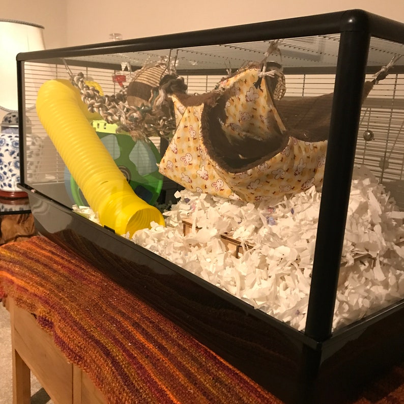 May include: A black glass hamster cage with a yellow tube, a brown and white hanging bed, and white shredded paper bedding.