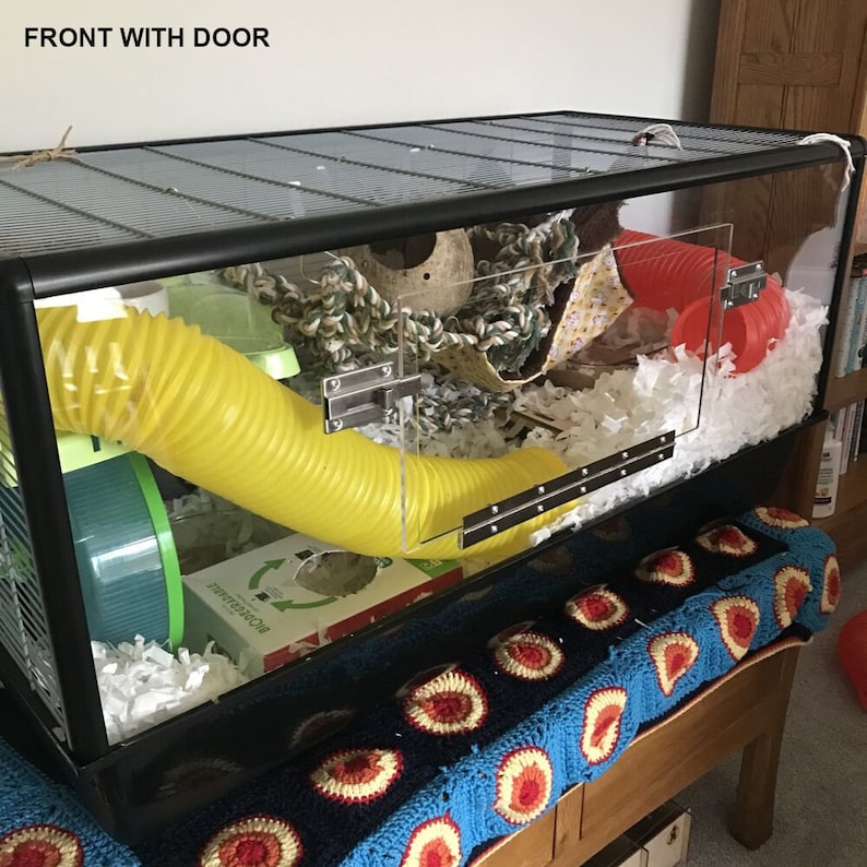 May include: A black and clear plastic hamster cage with a yellow tube, a red tube, a green wheel, and white bedding. The cage has a door on the front and a metal latch.