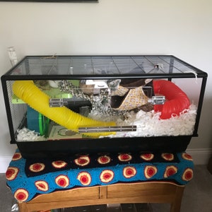 May include: A black wire cage with a yellow tube, a red tube, and white bedding. The cage is on a blue and white crocheted blanket.