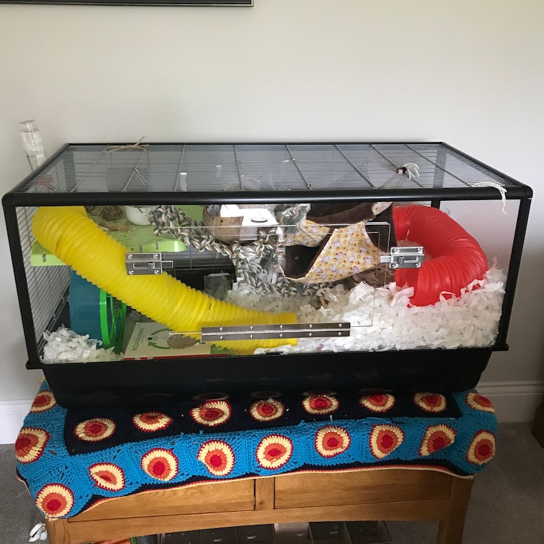 May include: A black wire cage with a yellow tube, a red tube, and white bedding. The cage is on a blue and white crocheted blanket.