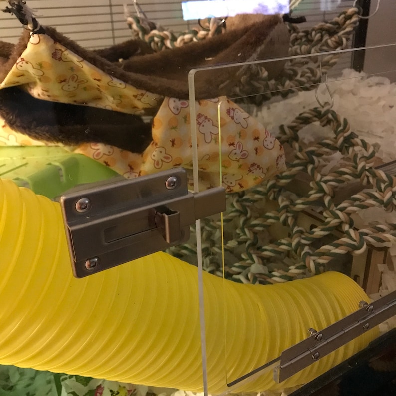 May include: A yellow plastic tube with a silver latch and a clear acrylic panel. The tube is part of a small animal cage, which also includes a brown and yellow fabric hammock and a green and white rope toy.