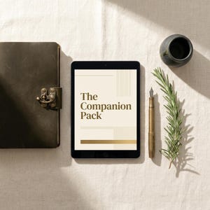 May include: An arrangement featuring a leather-bound journal with a clasp, a tablet displaying "The Companion Pack" in gold lettering, a brass fountain pen, a small ceramic vase, and a sprig of rosemary. The items are arranged on a neutral-toned surface.