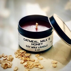 May include: A soy wax candle in a black tin with the lid slightly ajar. The candle is labeled "Oatmeal, Milk & Honey" and is 4 oz. Rolled oats are scattered around the candle.