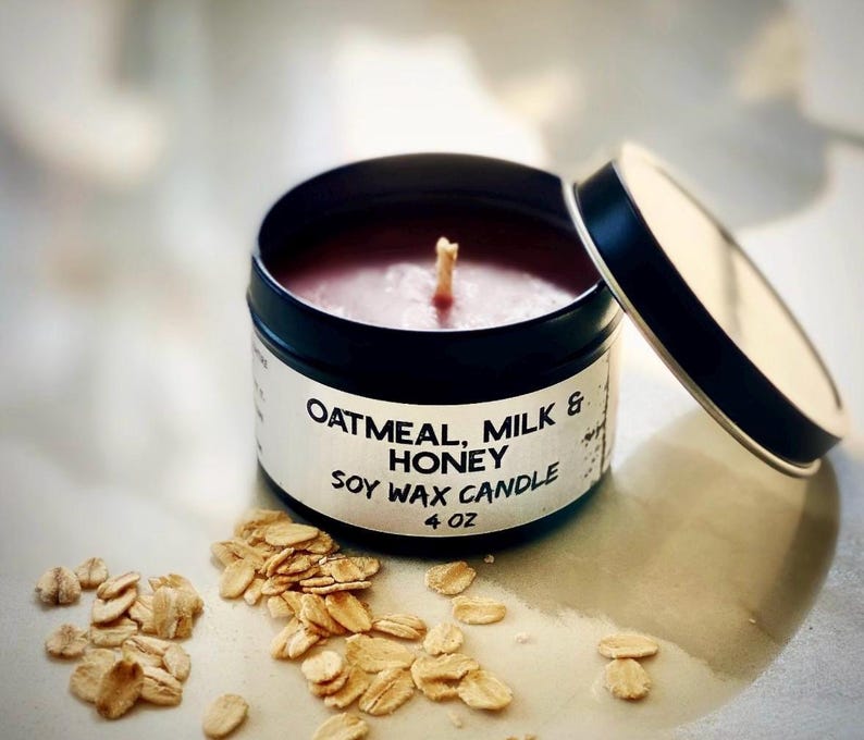 May include: A soy wax candle in a black tin with the lid slightly ajar. The candle is labeled "Oatmeal, Milk & Honey" and is 4 oz. Rolled oats are scattered around the candle.