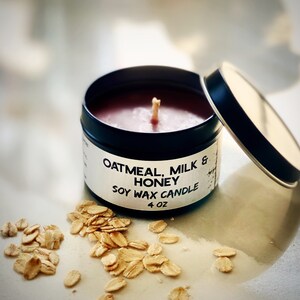 May include: A black tin candle with a partially open lid. The candle is filled with a reddish-brown wax and has a wooden wick. The label reads "Oatmeal, Milk & Honey Soy Wax Candle 4 oz." Rolled oats are scattered around the candle.