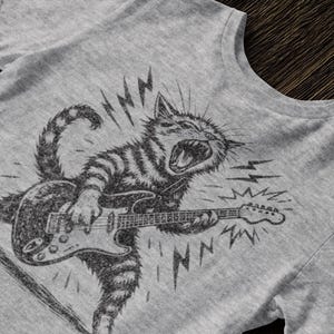 May include: Gray t-shirt featuring a black and white illustration of a cat playing an electric guitar. The cat is depicted with an open mouth and lightning bolts, suggesting a rock and roll theme. The design is printed on a gray t-shirt.