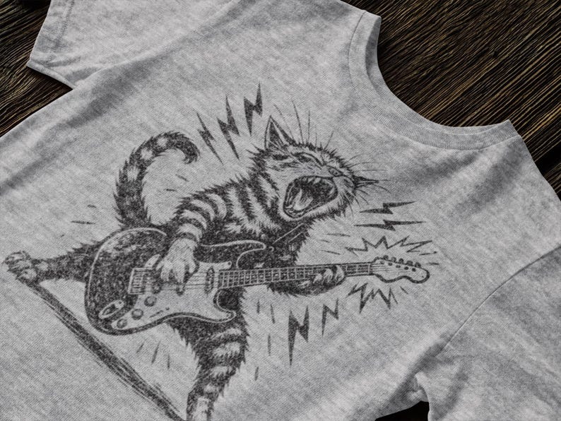 May include: Gray t-shirt featuring a black and white illustration of a cat playing an electric guitar. The cat is depicted with an open mouth and lightning bolts, suggesting a rock and roll theme. The design is printed on a gray t-shirt.