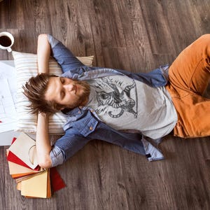 May include: A person lying on a wooden floor, wearing a gray graphic t-shirt, blue denim shirt, and orange pants. A pillow, coffee cup, and design samples are nearby. The person is resting with their eyes closed.