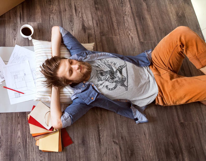 May include: A person lying on a wooden floor, wearing a gray graphic t-shirt, blue denim shirt, and orange pants. A pillow, coffee cup, and design samples are nearby. The person is resting with their eyes closed.