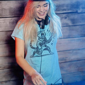 May include: A light blue t-shirt with a graphic of a tiger playing a guitar, worn with a black cap and headphones. The person is smiling and operating a DJ mixer. The background is a wooden wall.