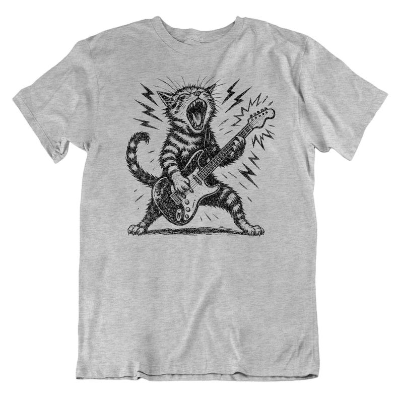 May include: A light gray t-shirt featuring a black and white illustration of a cat playing an electric guitar. The cat is roaring with lightning bolts surrounding it. The cat has striped fur and is in a dynamic pose.