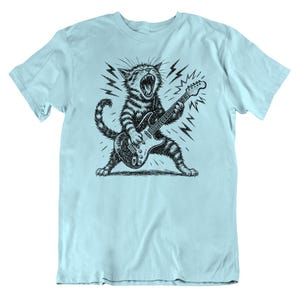 May include: Light blue t-shirt featuring a black and white illustration of a cat playing an electric guitar. The cat is depicted with its mouth open, surrounded by lightning bolt graphics, creating a rock and roll theme.
