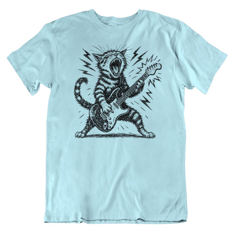 May include: Light blue t-shirt featuring a black and white illustration of a cat playing an electric guitar. The cat is depicted with its mouth open, surrounded by lightning bolt graphics, creating a rock and roll theme.