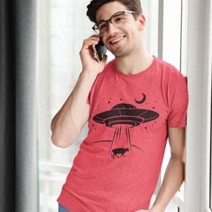 May include: A man wearing a red t-shirt with a black graphic of a UFO abducting a cow. He is talking on a cell phone and smiling.