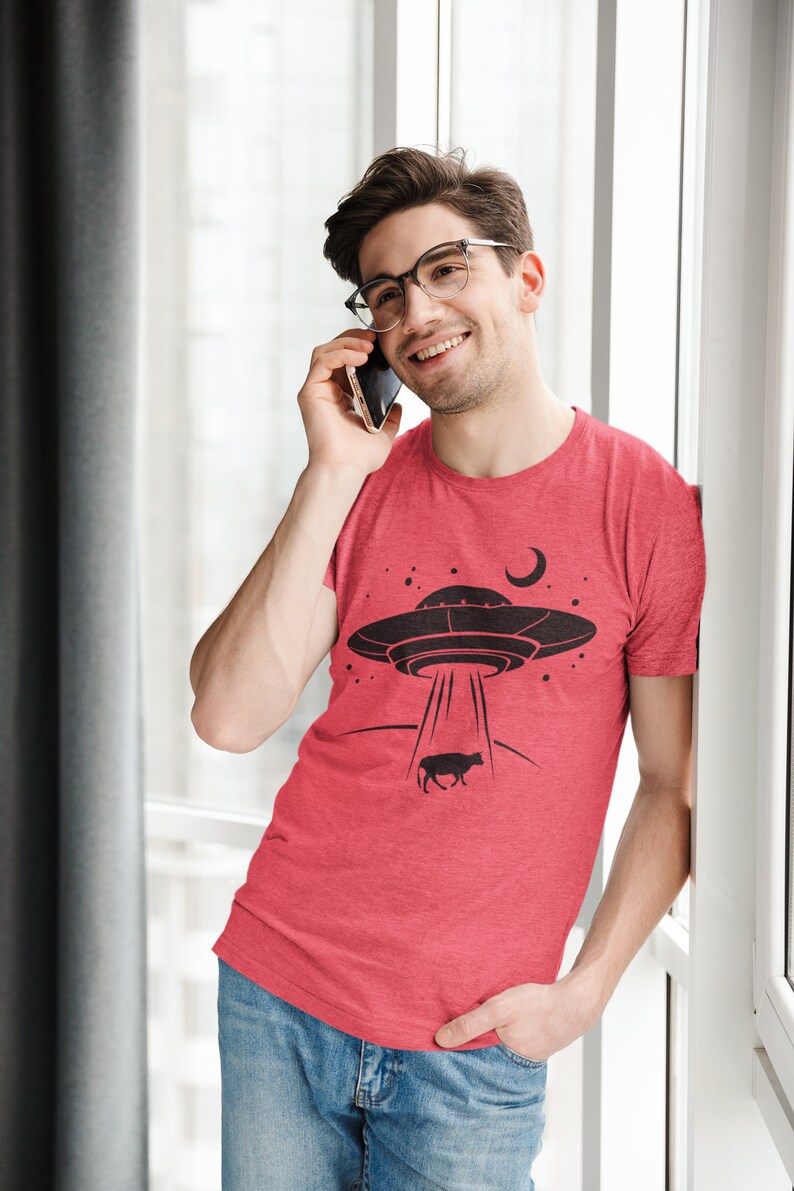 May include: A man wearing a red t-shirt with a black graphic of a UFO abducting a cow. He is talking on a cell phone and smiling.