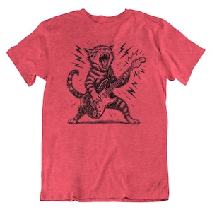 May include: Red t-shirt featuring a black and white illustration of a cat playing an electric guitar. The cat is roaring with lightning bolts in the background. The shirt is a crew neck and has short sleeves.