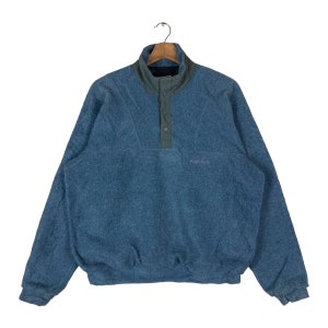 Może przedstawiać: Niebieski polarowy sweter z szarym kołnierzem i zapięciem na guziki. Top z długim rękawem ma teksturowany wygląd i małe logo na piersi. Ubranie wisi na drewnianym wieszaku.