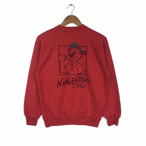Puede incluir: Sudadera roja con un dibujo de oso de dibujos animados con una gorra de béisbol. El dibujo está en un cuadrado con contorno negro. Debajo del dibujo está el texto "N.VALENTINO ITALY". La sudadera está colgada en una percha de madera.
