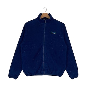 Może przedstawiać: Granatowa polarowa kurtka z pełnym zapięciem na zamek i stójką. Kurtka posiada małą prostokątną naszywkę z napisem "L.L.Bean" na lewej piersi. Kurtka wisi na drewnianym wieszaku.