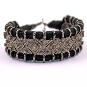 May include: A black and silver bracelet with a geometric design. The bracelet features a woven black band with silver-toned metal accents and diamond-shaped embellishments encrusted with small, clear crystals. The bracelet is designed to be worn around the wrist.