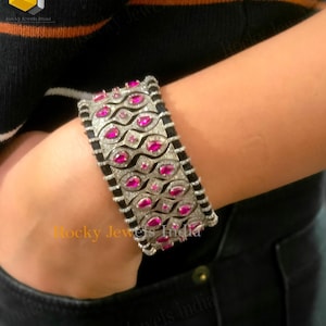 Può includere: Un braccialetto rigido in argento con una fascia nera e pietre preziose rosa. Il braccialetto è incastonato con un motivo ripetitivo di pietre preziose rosa di forma ovale e pietre preziose bianche più piccole. Il braccialetto viene indossato al polso.