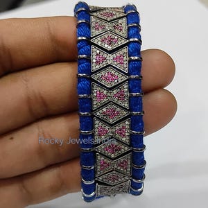 May include: A silver bracelet with a geometric design featuring pink and white gemstones. The bracelet is accented with bright blue cord. The text "Rocky Jewels India" is visible.
