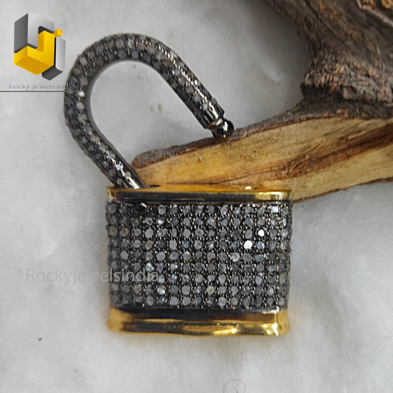 May include: A gold and silver padlock charm with a diamond-encrusted design. The padlock is open and the hasp is visible. The charm is set against a wooden background.
