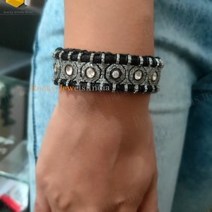 May include: A black and silver bracelet with a row of round white stones set in a silver frame. The bracelet is made of a black cord with silver accents. The text "Rocky Jewels India" is visible in the image.