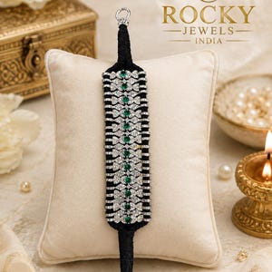 Pode incluir: Uma pulseira preta e prateada com detalhes em verde esmeralda. A pulseira apresenta uma série de desenhos em forma de laço, alternados com fileiras de pedras brilhantes. É apresentada numa almofada creme, com o logótipo "Rocky Jewels India" no fundo.