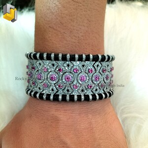 May include: A wide silver bracelet with a geometric design, featuring pink gemstones and small clear stones. The bracelet has a black band on each side. The bracelet is approximately 2 inches wide.