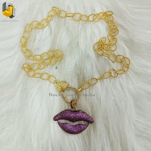 May include: A gold chain necklace with a purple lip pendant encrusted with sparkling stones. The necklace features a unique clasp design. RockJewIndia is printed on the image.
