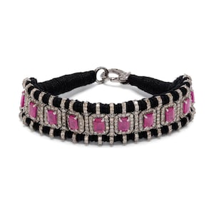 May include: A black cord choker necklace with a silver clasp. The necklace features a row of rectangular, pink gemstones surrounded by small, clear stones. The gemstones are set in a silver metal frame.
