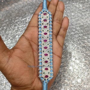 May include: A light blue bracelet with a central rectangular design. The design features alternating red and green gemstones, surrounded by smaller clear stones. The bracelet is held together with a light blue cord and a silver clasp.